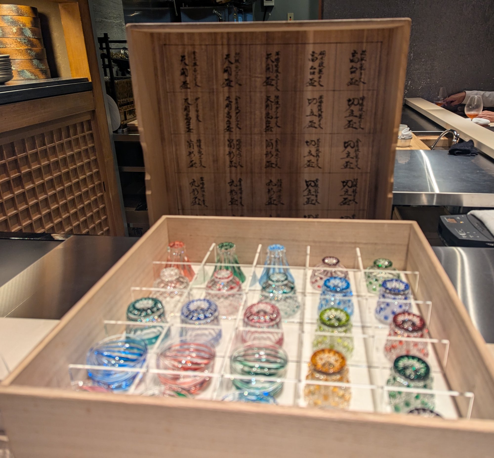 A wooden box containing an array of Horiguchi kiriko cut-glass sake cups, with handwritten notes on the inside of the lid